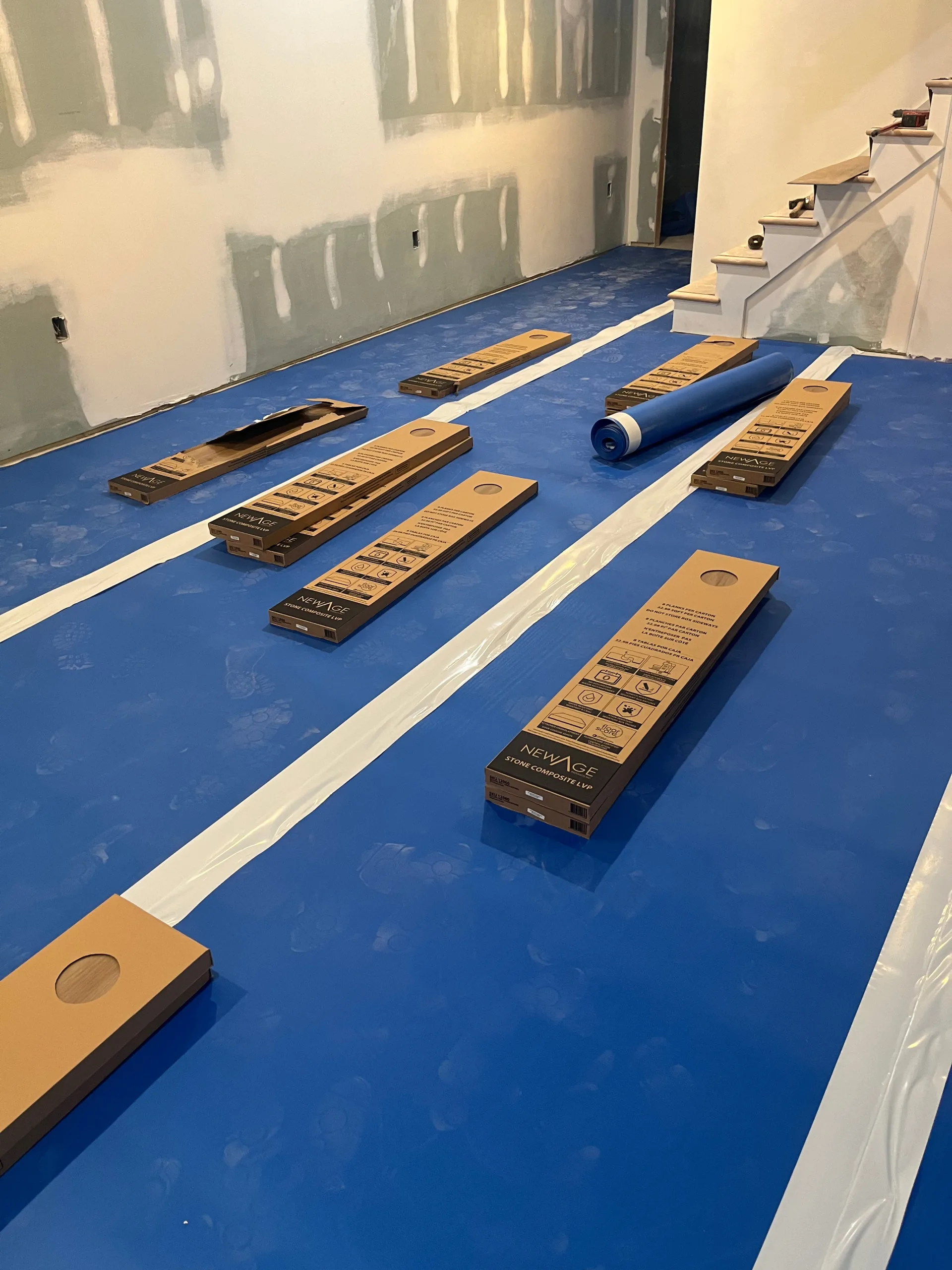 Overview of a basement room prepped with blue protective film and multiple staged boxes of flooring for a comprehensive composite vinyl plank installation.
