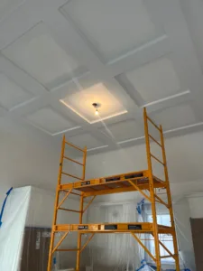 Looking up at a beautifully detailed, painted multi-panel Custom Coffered Ceiling Finishing, with a yellow scaffolding tower set up below to provide access for painters.