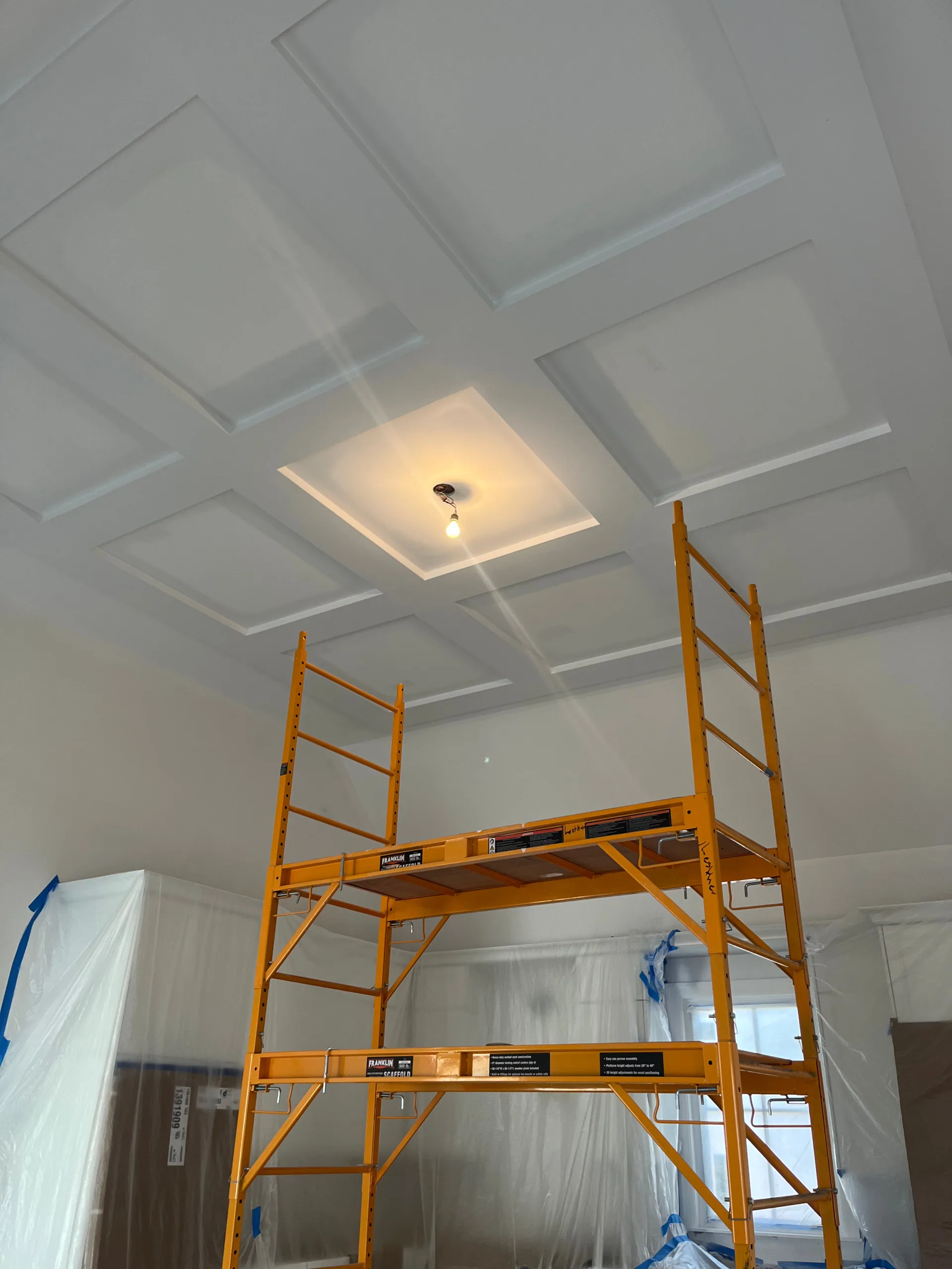Looking up at a beautifully detailed, painted multi-panel Custom Coffered Ceiling Finishing, with a yellow scaffolding tower set up below to provide access for painters.