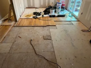 Hardwood floor installation in progress showing exposed plywood subfloor, pneumatic tools, and partial wood layout.