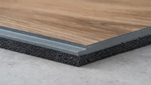 Close-up photo of a vinyl flooring section showing its multi-layer structure. The top surface features a brown wood-grain finish over a solid gray layer and a thick, porous black foam base (acoustic underlayment) for sound absorption.