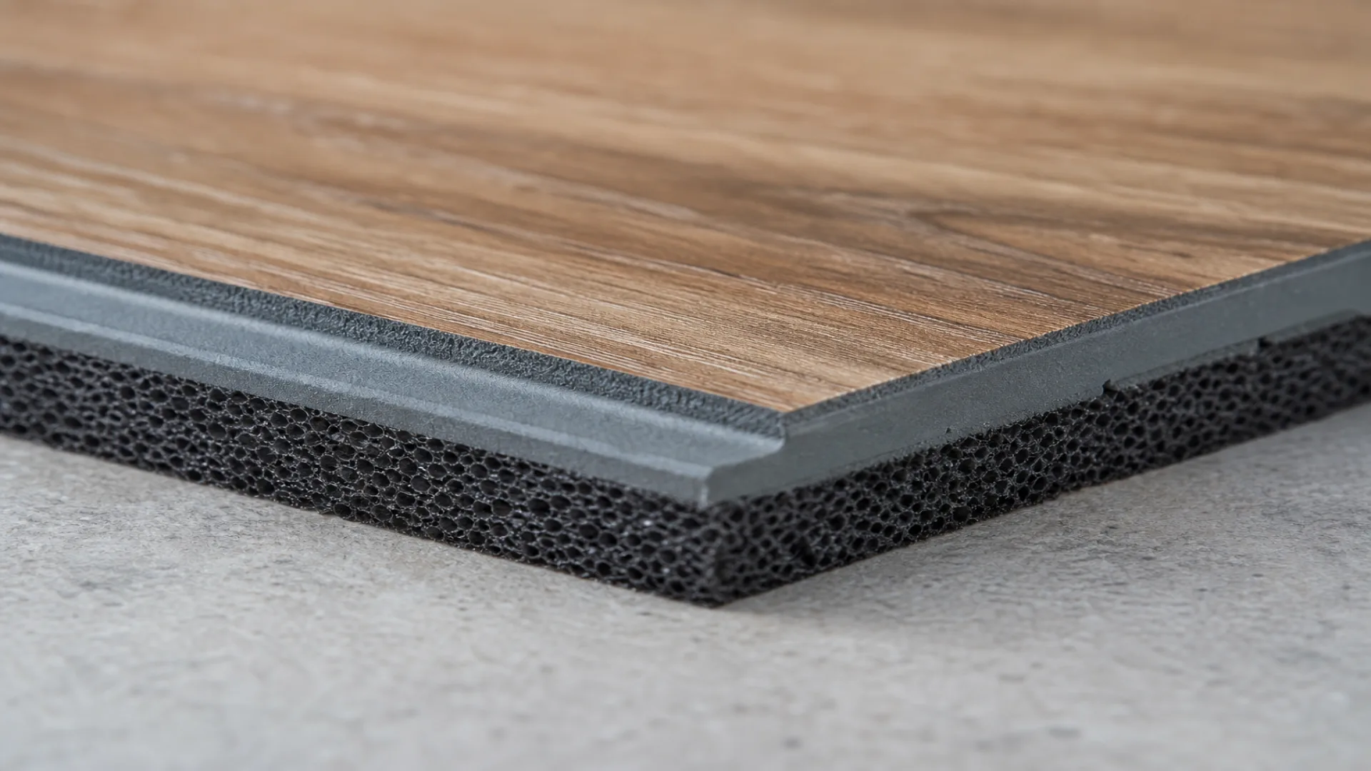 Close-up photo of a vinyl flooring section showing its multi-layer structure. The top surface features a brown wood-grain finish over a solid gray layer and a thick, porous black foam base (acoustic underlayment) for sound absorption.
